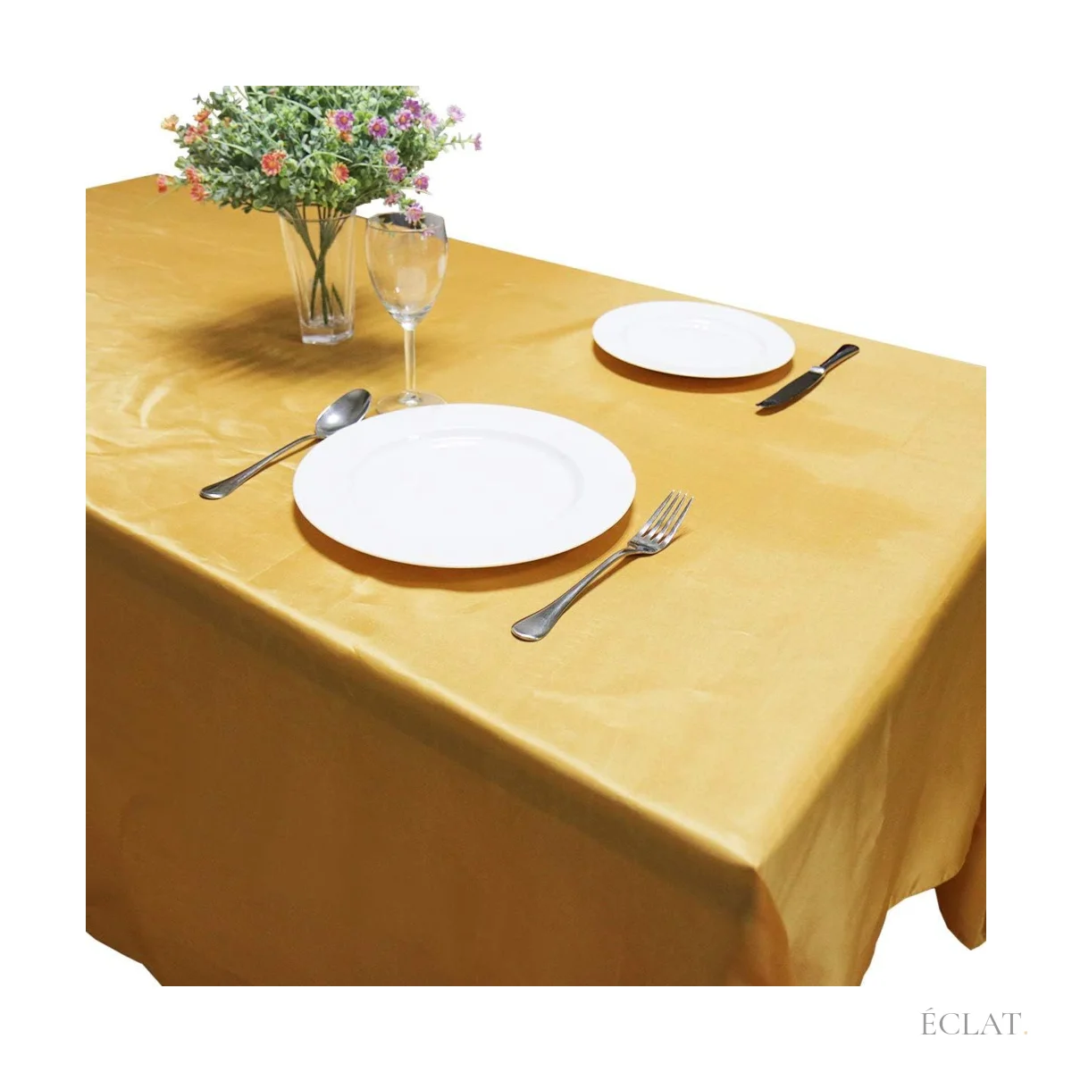 Nappe satin or dressée sur une table rectangulaire avec assiettes blanches et bouquet de fleurs